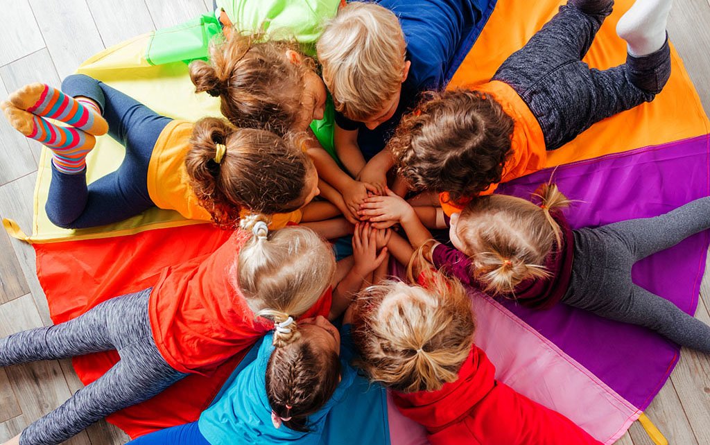 Lovely children laying on multicolor canopy in circle. Team building game for daycare children in colorful t-shirts. Top view kids on wooden floor.